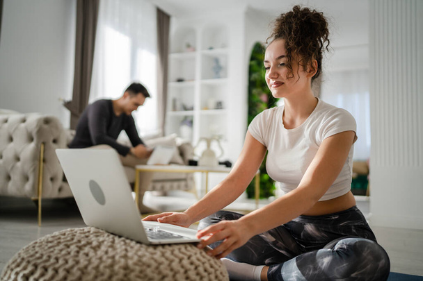Woman young adult caucasian female sitting on the floor at home working on laptop computer browse internet online while her husband or boyfriend is sitting in the background leisure family concept - Фото, изображение
