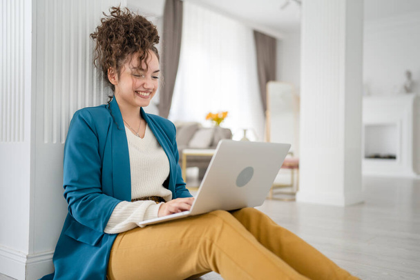 Une femme jeune adulte caucasienne assise sur le sol à la maison avec ordinateur portable travaillant et regardant vers le côté heureux sourire brunette indépendante avec des cheveux bouclés vie domestique copie espace - Photo, image