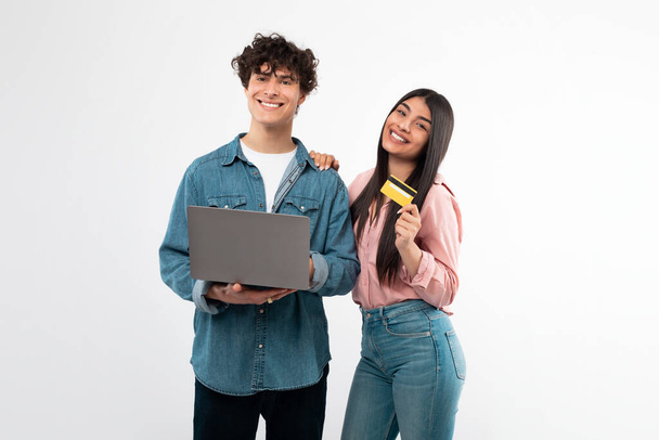 E-Commerce-Angebot. Fröhliches junges Paar, das mit Laptop und Kreditkarte online einkauft, vor weißem Studiohintergrund posiert und in die Kamera lächelt. Einfaches Zahlungskonzept - Foto, Bild