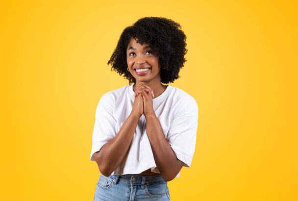 Χαρούμενη νεαρή Αφροαμερικανή σγουρή γυναίκα που κάνει κίνηση προσευχής με τα χέρια, ονειρεύεται, κάνει μια ευχή, απομονωμένη στο κίτρινο φόντο του στούντιο. Ζήτα, ελπίδα και πίστη, ευγνωμοσύνη και αγάπη - Φωτογραφία, εικόνα
