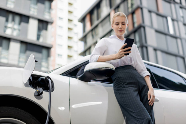 Closeup progressive businesswoman talking on smartphone, leaning electric car at charging station with residential condo apartment in background. Eco friendly rechargeable EV car concept. - Fotó, kép