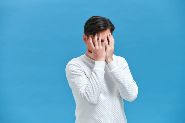 Depressed and frustrated. Frustrated young man touching head with hand while standing against blue background - 写真・画像