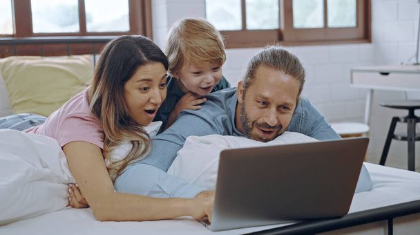 Famiglia caucasica di tre persone che usano il computer portatile mentre sono sdraiati sul letto insieme, navigano in internet o guardano film. - Foto, immagini