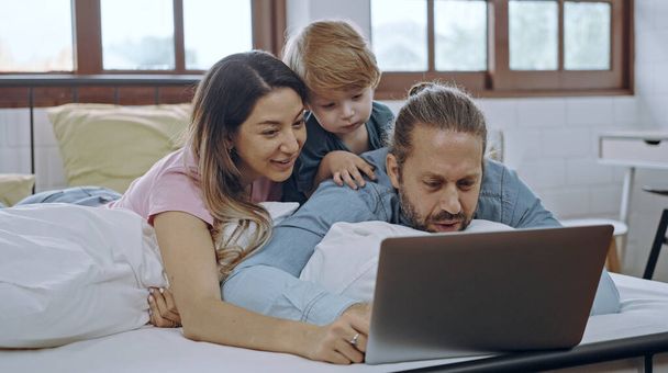 Famiglia caucasica di tre persone che usano il computer portatile mentre sono sdraiati sul letto insieme, navigano in internet o guardano film. - Foto, immagini