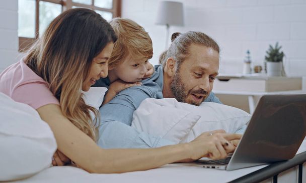 Famille caucasienne de trois personnes utilisant un ordinateur portable tout en étant couchés ensemble, naviguant sur Internet ou regardant un film. - Photo, image