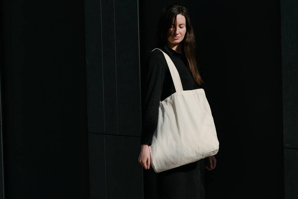 Woman holding white textile eco bag against urban city background. Ecology or environment protection concept. White eco bag for mock up. - Фото, зображення