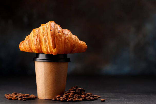Aromatic coffee in a paper cup paired with a flaky croissant. With copy space - Photo, Image