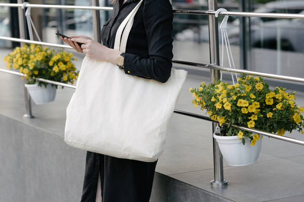 Kobieta trzyma białą tekstylne eko worek przed miejskim tle miasta. Ekologia lub koncepcja ochrony środowiska. Biała torba eko do makiety. - Zdjęcie, obraz