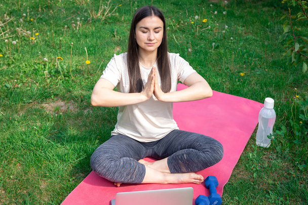 A young woman meditates after a workout, sitting in front of a laptop on a mat in a green park. - Photo, Image