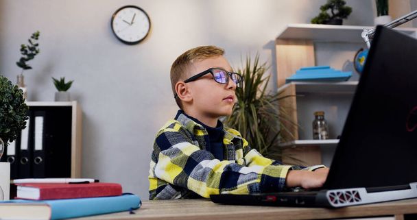 Portrait of attractive teen smart boy in eyeglasses which sitting at his workplace and doing homework using computer - Фото, зображення