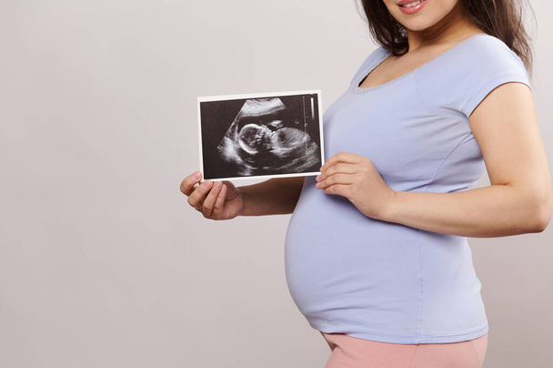 Крупный план большого живота улыбающейся беременной женщины, будущей матери, держащей в руках ультразвуковое изображение своего будущего ребенка, на изолированном студийном фоне. Беременность. Роды. Ожидание. Материнство - Фото, изображение