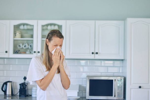 Allergieën, toiletpapier en een vrouw die haar neus snuit in de keuken voor een verkoudheid, griep of niezen bij haar thuis. Ziekte, virus en jonge vrouwelijke persoon met weefsel voor sinus, hooikoorts of covid in appartement - Foto, afbeelding