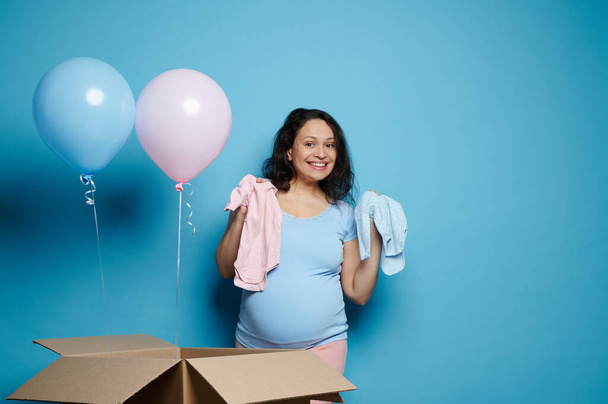 Happy pregnant woman with big belly, in 2nd trimester of her pregnancy, showing a blue and pink newborn bodysuit, expressing amazement at gender reveal party, baby shower, isolated on blue background - Foto, Bild