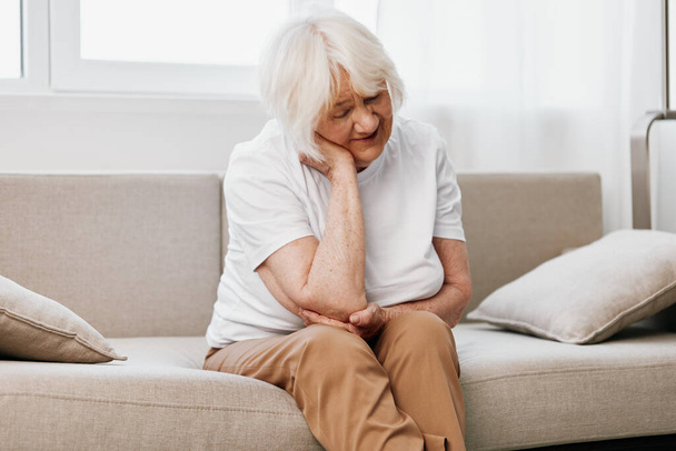 Пожилая женщина, сильная боль в шее, сидя на диване, проблемы со здоровьем в преклонном возрасте, плохое качество жизни. Высокое качество фото - Фото, изображение
