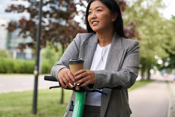 Chinese woman standing on electric scooter in business outfit and holding a cup of coffee - Foto, afbeelding
