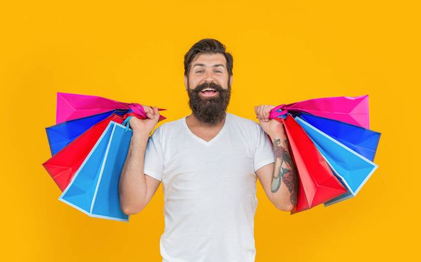 smiling shopaholic man at shopping in studio. shopaholic man at shopping on background. photo of shopaholic man at shopping hold bags. shopaholic man at shopping isolated on yellow. - 写真・画像
