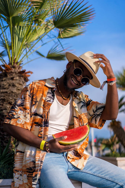 Portret van een zwarte etnische man die geniet van de zomervakantie op het strand, met een hoed en een watermeloen op - Foto, afbeelding
