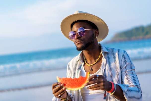 Portrait of a black ethnic man enjoy summer vacation at the beach eating a watermelon wearing a sunshade and sunglasses - Valokuva, kuva