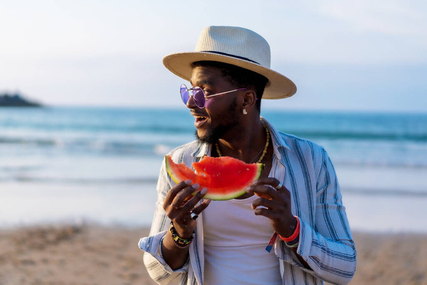 Black ethnic man enjoy summer vacation on the beach eating a watermelon by the sea - Photo, Image