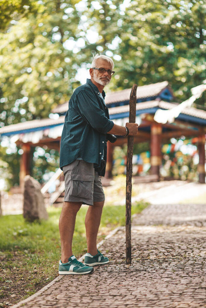 Sopalı ve sırtı ağrıyan yaşlı bir adam parkta yürüyor. 60 'larda yürüyüş. - Fotoğraf, Görsel