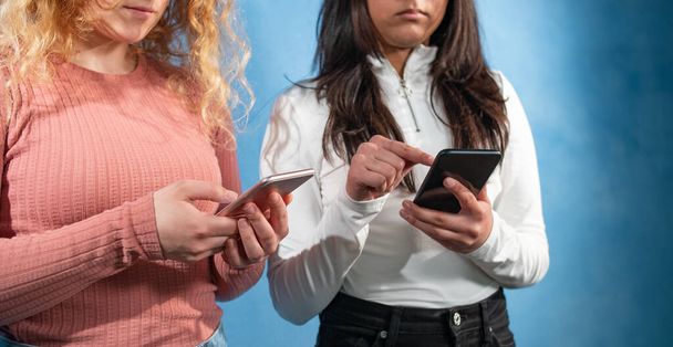 Two teenager typing on their phones. Two young women stands at a blue background. They are in thought and types on their mobile phones. - Foto, Imagem