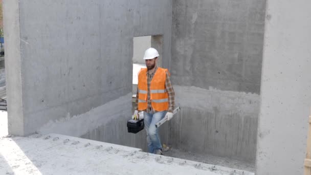 Stavební opravář, předák v ochranné přilbě a vestě stojí na pracovišti v budově a drží pravítko a skříňku s nářadím v rukou - Záběry, video