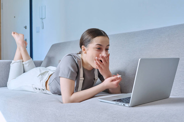 Teen girl at home lying on couch, talking laughing looking at laptop webcam, having video chat call conference. Technology for learning leisure communication, modern lifestyle, adolescence concept - Фото, зображення