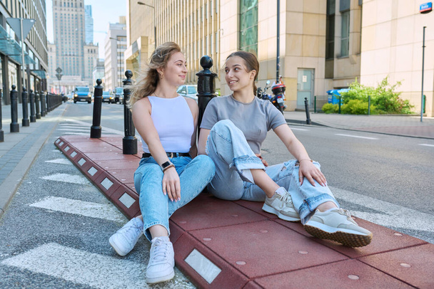 Two smiling teenage female students looking at camera, modern urban style, buildings background. Fashionable girls sitting on sidewalk near road. Youth, fashion, style, city, lifestyle concept - Fotoğraf, Görsel