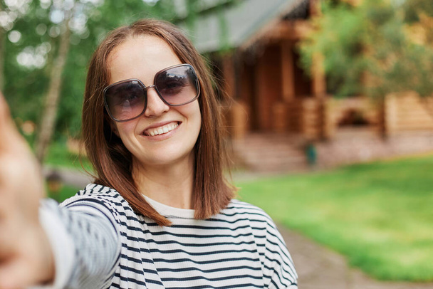 Smiling young dark haired girl making selfie photo in park with wooden house on background expressing positive emotions, POV, woman broadcasting livestream, having video call. - Foto, afbeelding