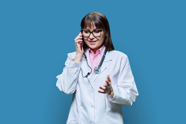 Mature female doctor in white lab coat talking on phone, on blue studio background. Medicine, service, occupation, healthcare, health care, treatment concept - Foto, Imagem