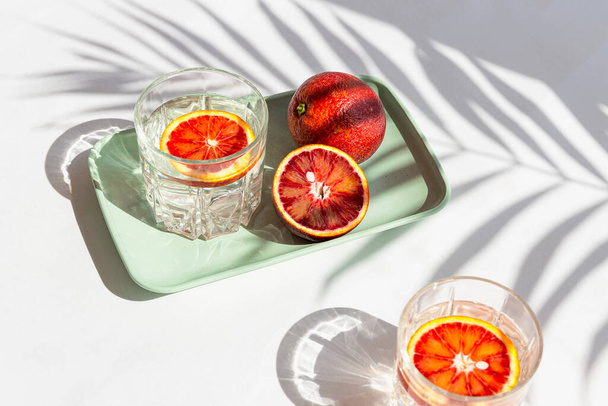 Kreative Komposition aus halb blutiger roter Orange auf einem Tablett und zwei Gläsern mit Cocktail auf weißem Hintergrund mit Palmblattschatten. Sommerfrische und Party-Konzept. Minimaler Stil. - Foto, Bild
