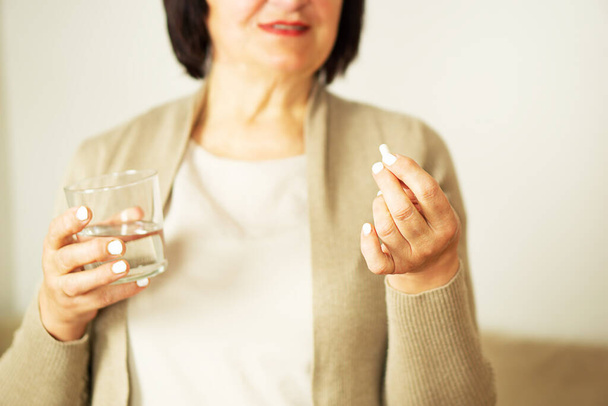 Nyugdíjas nő alkalmi ruhában otthon, tablettával és üvegvízzel a kezében. Egészséges, középkorú nő, kezében egy pohár vízzel és a gyógyszereivel. Boldog felnőtt nő vitamintablettát szed. - Fotó, kép