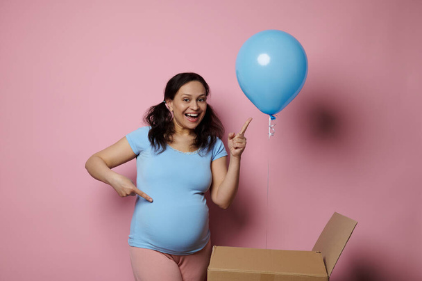 Expectant future mom, pregnant woman smiling and pointing finger at blue balloon, shes expecting a baby boy, isolated on pink background. Gender reveal party concept. Pregnancy. Maternity. Motherhood - Photo, Image