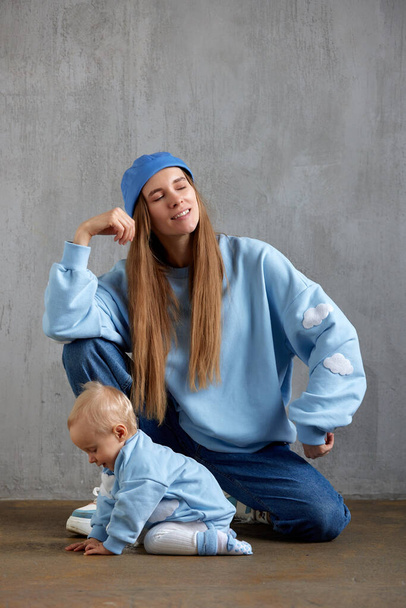 Young happy mom and her cute baby are sitting on the floor, chatting and playing. Mom and baby wear similar blue clothes. The concept of happy motherhood and childhood. Studio shot, gray background - Foto, imagen