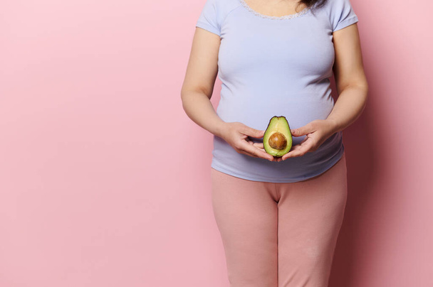 Midsection of pregnant expectant woman holding a half of avocado fruit at the level of her big belly, isolated over pink background. Pregnancy. Prenatal and maternity lifestyle. Womens health concept - 写真・画像