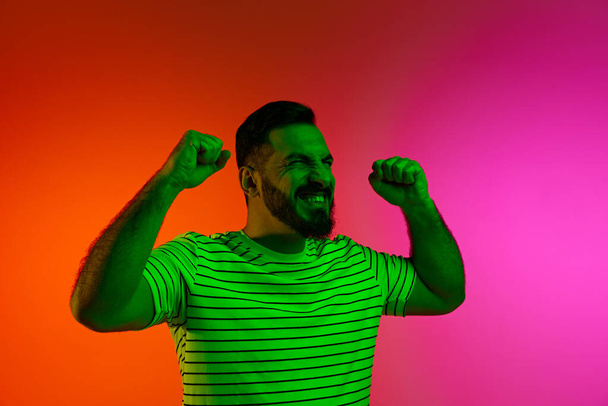 Portrait of happy bearded man posing with gesture of win and success against gradient studio background in neon light. Concept of human emotions, facial expression, lifestyle, business, promotion - Fotografie, Obrázek