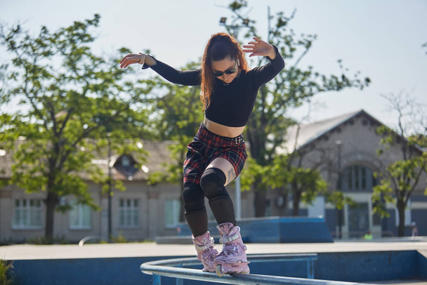 Cool young roller blader female grinding on a rail in a skatepark. White female person wearing aggressive inline skates performing a soul grind trick on a handrail - Fotó, kép