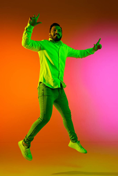 Full-length portrait of handsome bearded man in casual clothes jumping against gradient studio background in neon light. Concept of human emotions, facial expression, lifestyle, fashion - Fotó, kép