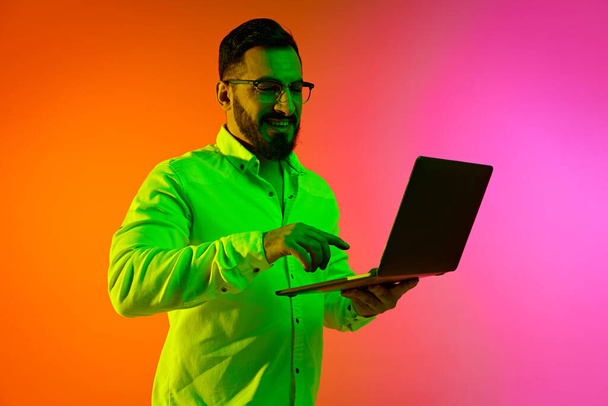 Bearded confident man in glasses working with laptop against gradient studio background in neon light. Concept of human emotions, facial expression, lifestyle, business, success, online shopping - Photo, Image