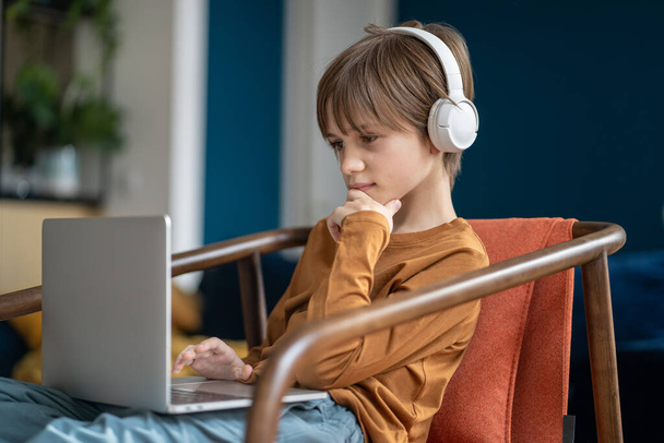 Задумчивый мальчик сидит на кресле и сидит дома, изучая ноутбук. Заинтересованный школьник набирает текст на компьютере, пользуется интернетом, дистанционным обучением, играет в онлайн видеоигры.  - Фото, изображение