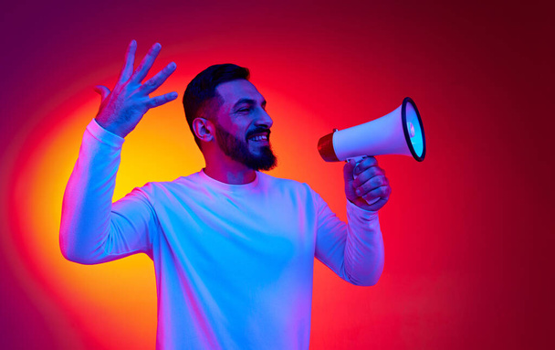 Portrait of excited, happy, smiling man in striped shirt shouting in megaphone against gradient red studio background in neon light. Concept of human emotions, facial expression, lifestyle - Foto, Imagen