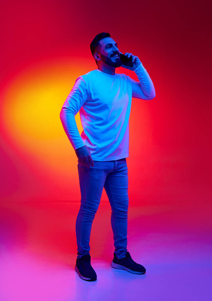 Portrait of handsome, smiling, bearded man in casual clothes talking on mobile phone against gradient studio background in neon light. Concept of human emotions, facial expression, lifestyle - Valokuva, kuva
