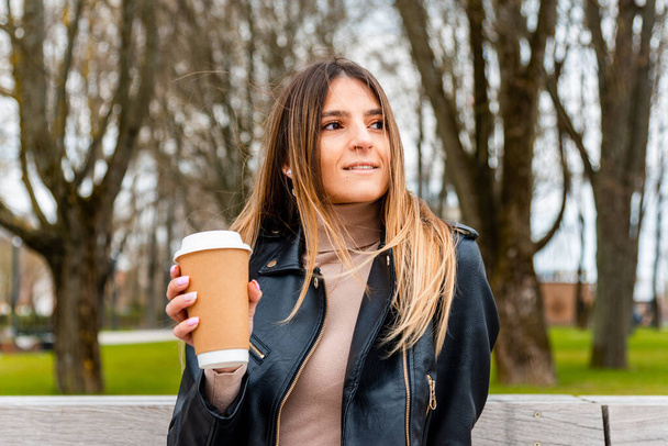 Радостная молодая белая женщина с чашкой кофе на бумаге расслабляется на скамейке в весеннем парке.. - Фото, изображение