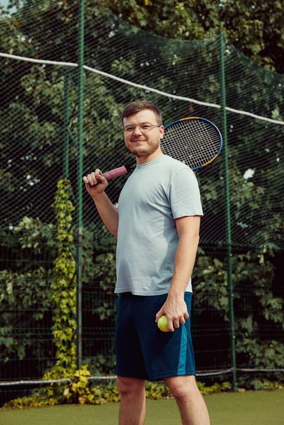 Πορτρέτο του νεαρού τενίστα στα αθλητικά με τη ρακέτα και την μπάλα στο χέρι να στέκεται στο γήπεδο του τένις - Φωτογραφία, εικόνα