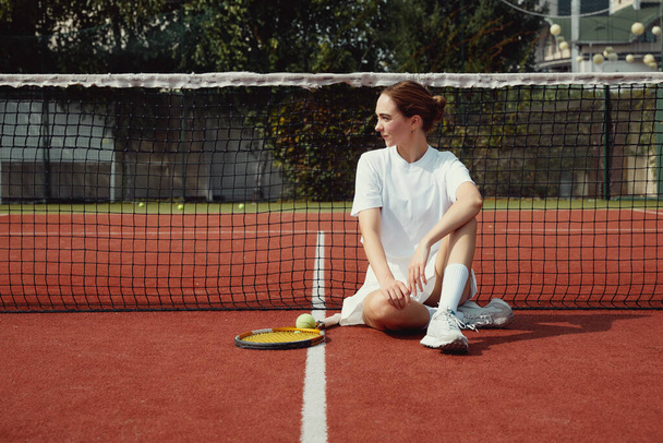Tenis raketi giyen beyaz elbiseli genç bir kadın tenis kortunda oturuyor ve sağa bakıyor. - Fotoğraf, Görsel
