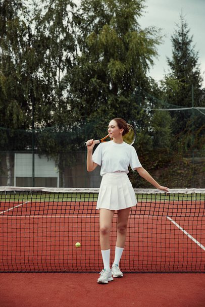 Vertikální fotografie módní dívky v bílém oblečení držící tenisovou raketu za hlavou, pózující na tenisové síti na hřišti. Sportovní móda - Fotografie, Obrázek