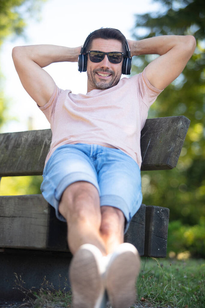 relaxed man listening music using headphones outdoors on bench - Foto, Imagen