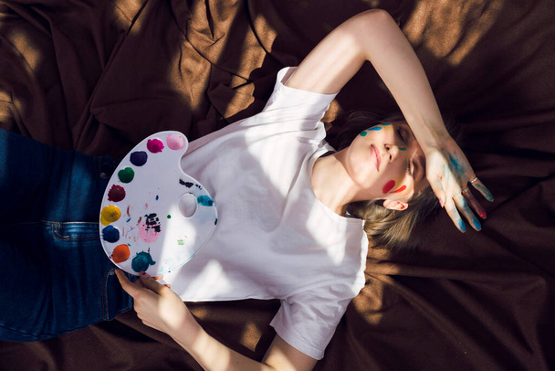 Roztomilé uvolněné blondýny dívka, malíř nebo umělec ležící na hnědé látky v pohodlné dílně, jíst pizzu a malování pod slunečními paprsky padající na ni. - Fotografie, Obrázek