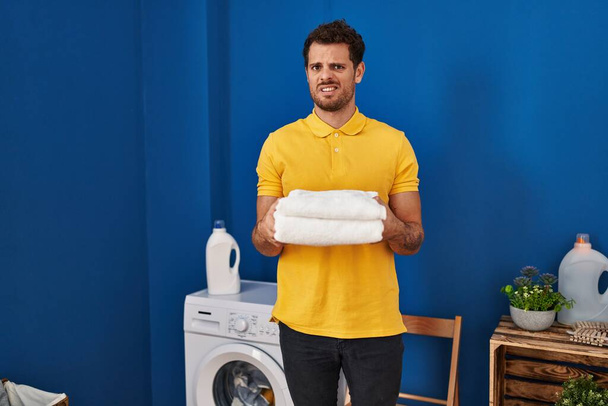 Jonge Spaanse man met schone handdoeken in de wasruimte, onwetend en verwarde uitdrukking. twijfelconcept.  - Foto, afbeelding
