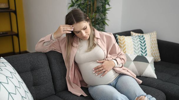 Молодая беременная женщина трогает живот с беспокойным выражением лица дома - Фото, изображение